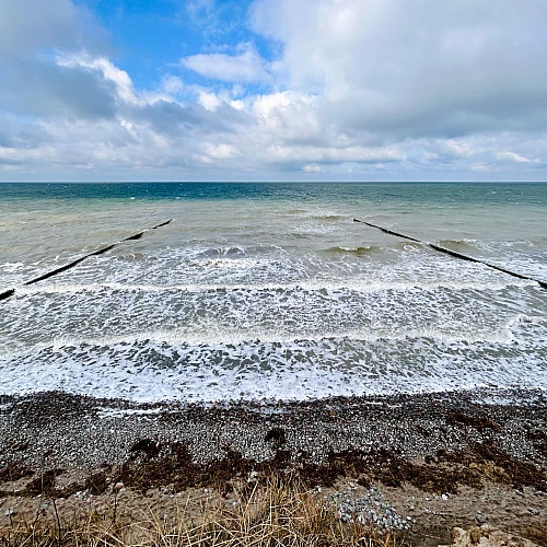 Im Vordergrund das Hohe Ufer, Richtung Horizont das stürmische Meer. Im oberen Drittel ein wolkenverhangener blauer Himmel.