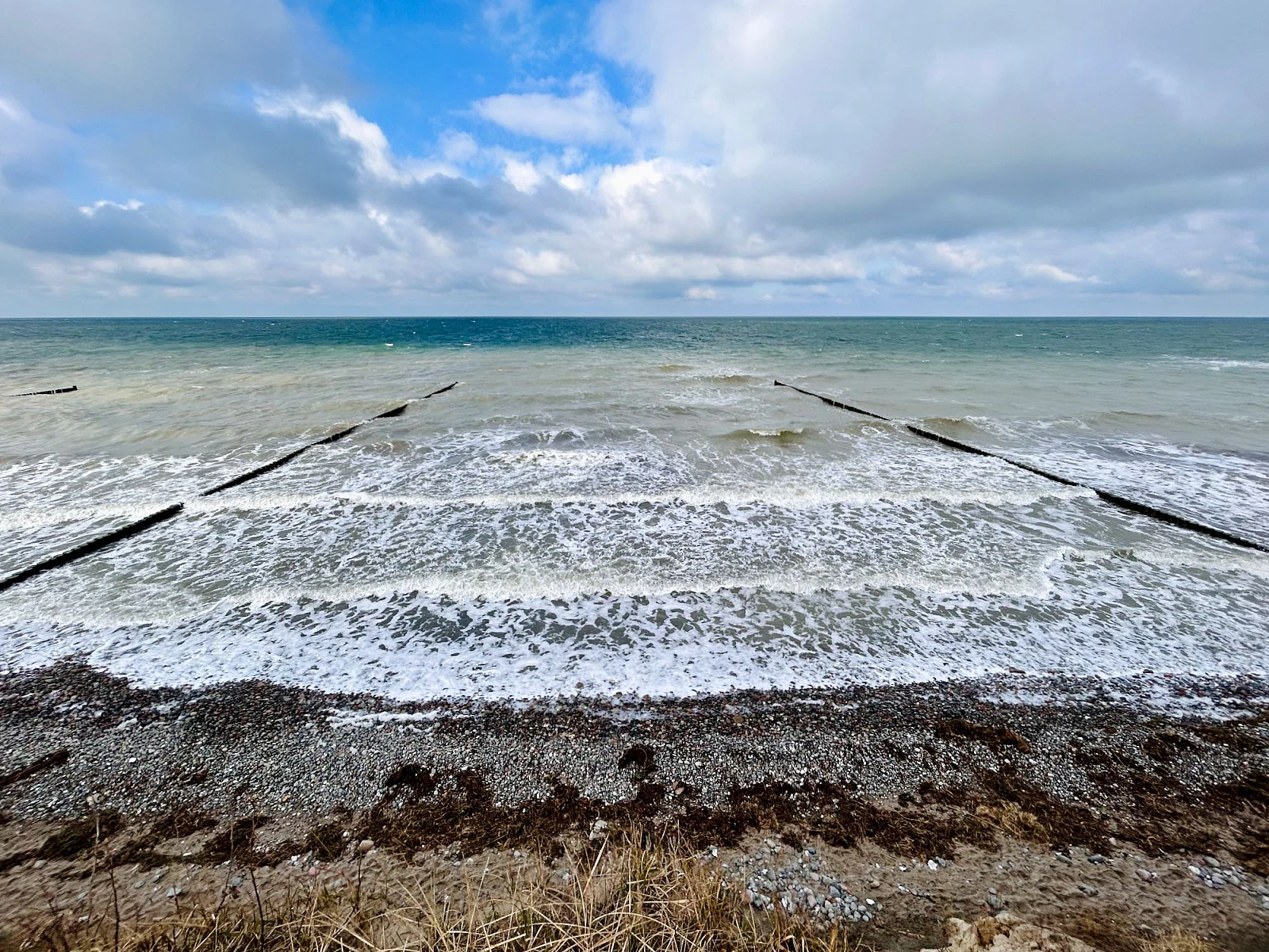 Im Vordergrund das Hohe Ufer, Richtung Horizont das stürmische Meer. Im oberen Drittel ein wolkenverhangener blauer Himmel.