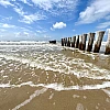 Rechts zieht sich vom Horizon zum Bildrand eine Reihe Holzbuhnen. Das Bild ist horizontal geteilt in ein Drittel Himmel, zwei Drittel Meer mit leichter auslaufender Brandung.