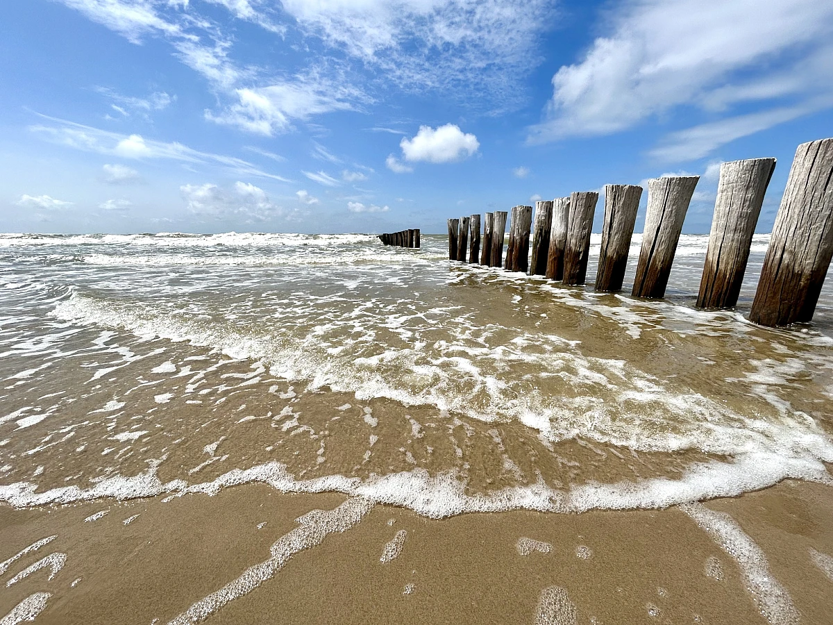 Im oberen Drittel des Bildes Himmel mit weißen Wolken, die folgenden zwei Drittel: nasser Strand auf dem sich der Himmel spiegelt.