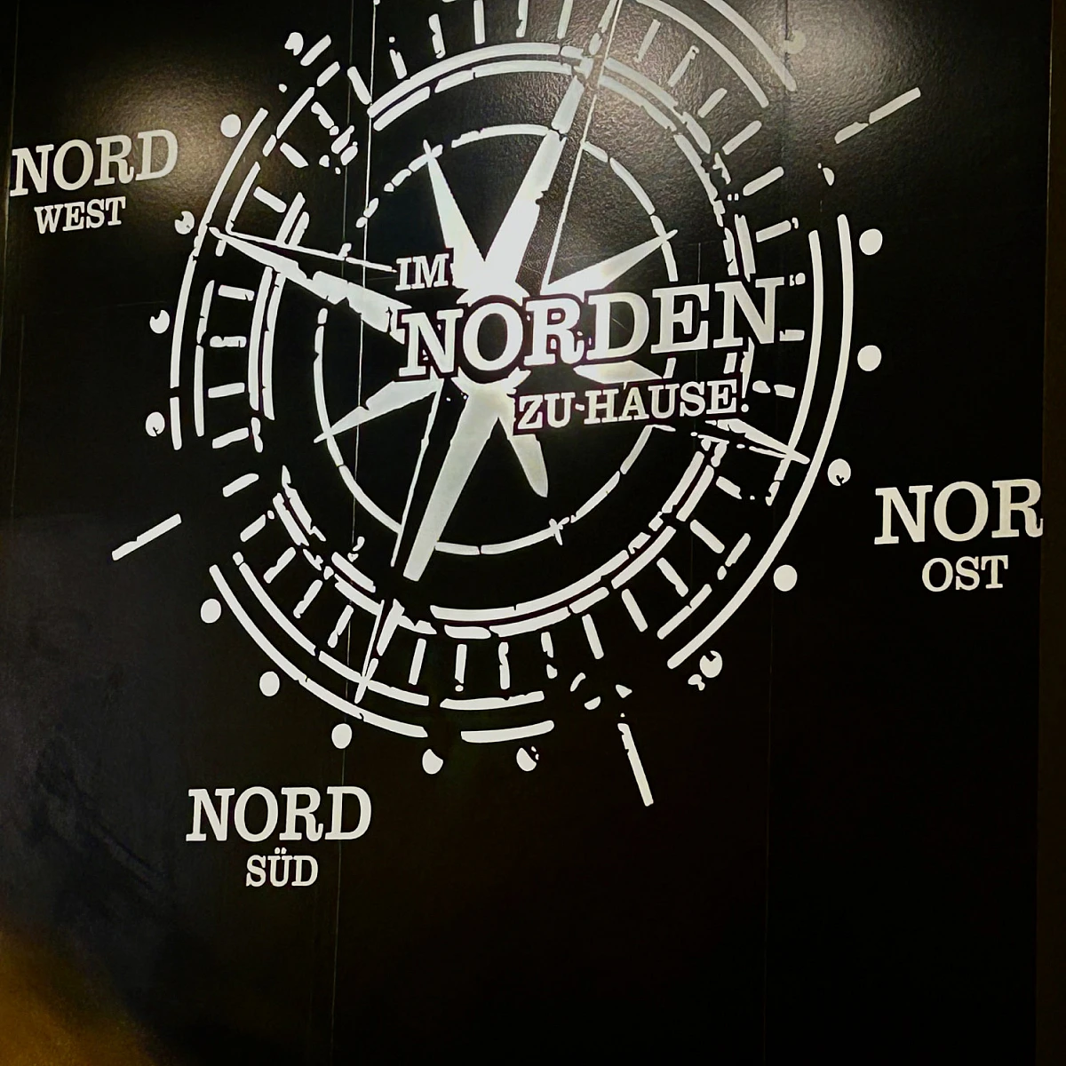 Foto eines Wandbildes. Auf schwarzem Grund ist ein Kompass zu sehen, dessen Richtungen Nord Nord, Nord Ost, Nord Süd und Nord West sind. Im Zentrum steht: Im Norden zu Hause