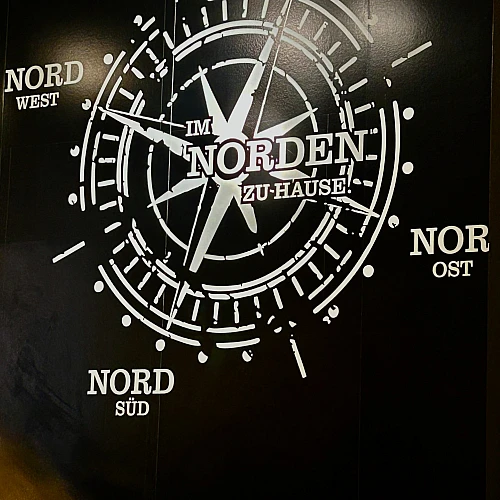 Foto eines Wandbildes. Auf schwarzem Grund ist ein Kompass zu sehen, dessen Richtungen Nord Nord, Nord Ost, Nord Süd und Nord West sind. Im Zentrum steht: Im Norden zu Hause