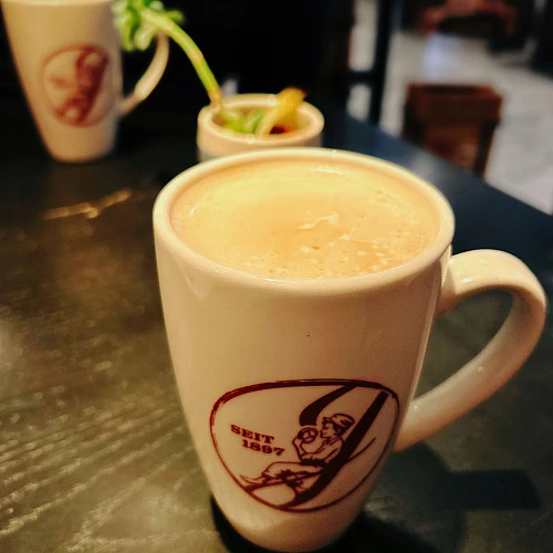 Auf einem schwarzen Holztisch stehen eine gefüllte Kaffeetasse im Vordergrund, eine im Hintergrund. Dazwischen eine kleine Pflanze.