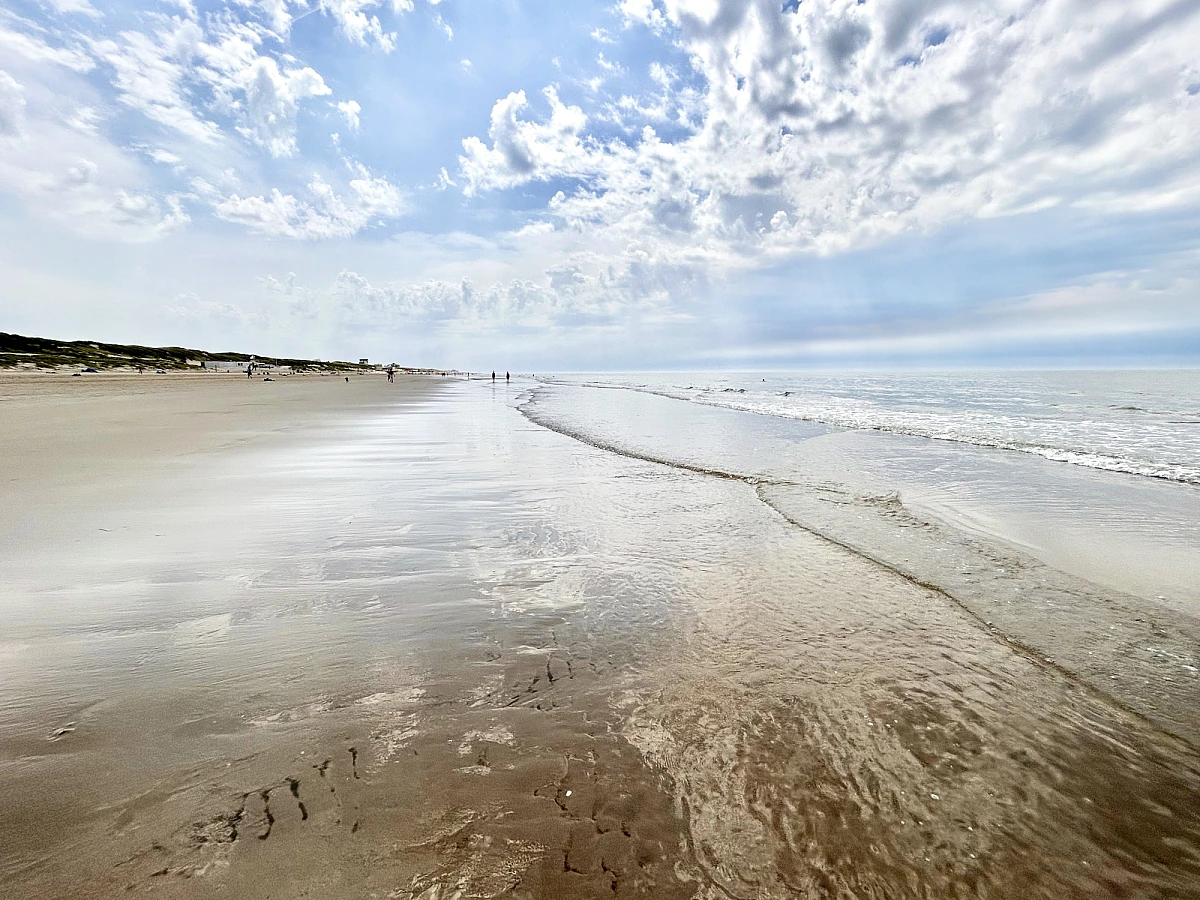 Im oberen Drittel des Bildes Himmel mit weißen Wolken, die folgenden zwei Drittel: nasser Strand auf dem sich der Himmel spiegelt.