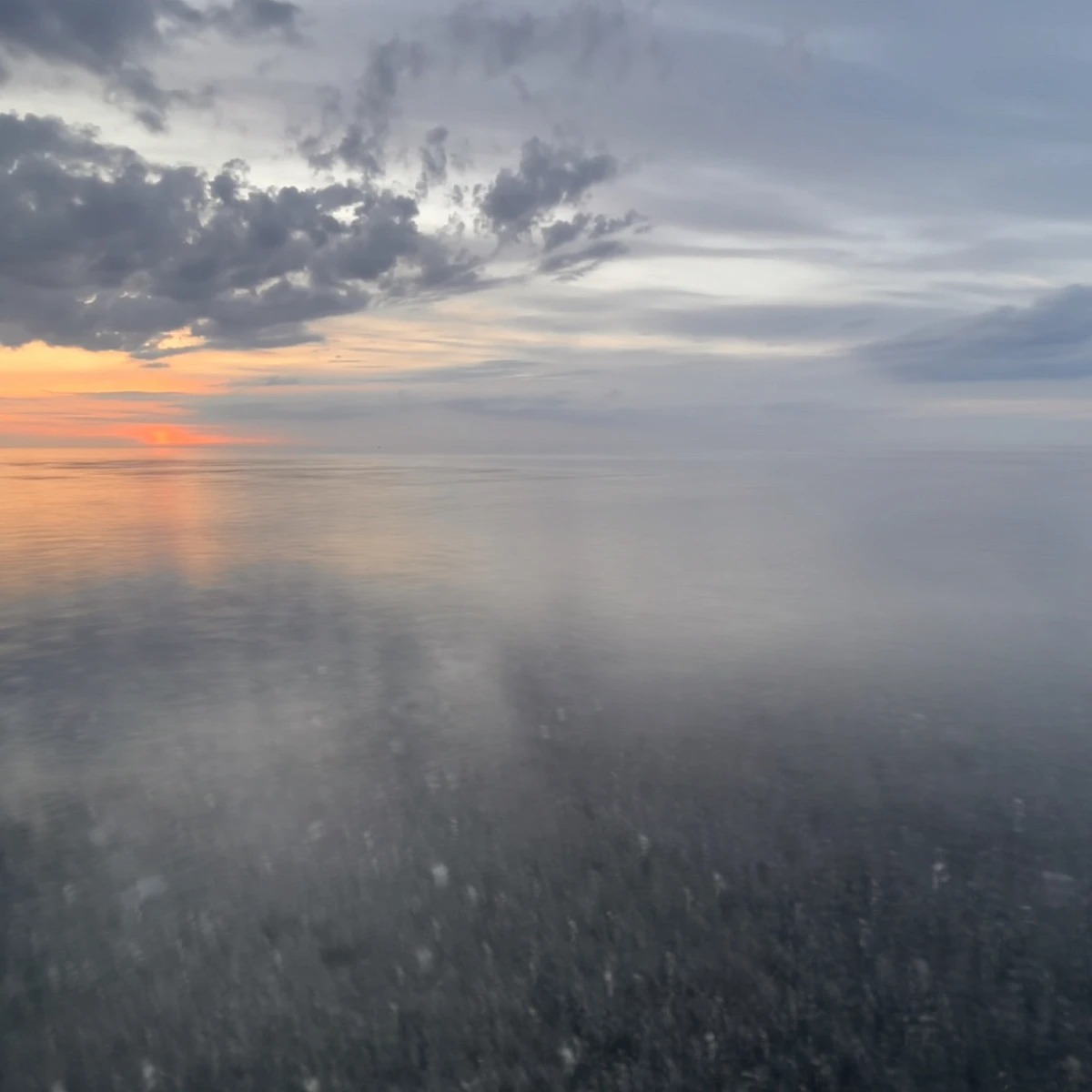 Sonnenuntergang. Das Bild ist gedrittelt. Zwei Drittel nimmt das durch die Langzeitbelichtung verschwommene Meer ein, ein Drittel die leicht orangene Sonne und bläulich graue Wolken am Himmel.