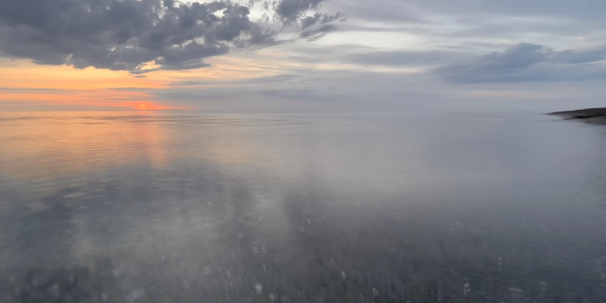 Sonnenuntergang. Das Bild ist gedrittelt. Zwei Drittel nimmt das durch die Langzeitbelichtung verschwommene Meer ein, ein Drittel die leicht orangene Sonne und bläulich graue Wolken am Himmel.