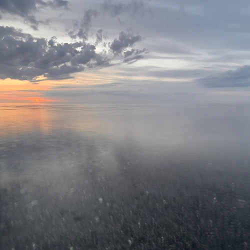 Sonnenuntergang. Das Bild ist gedrittelt. Zwei Drittel nimmt das durch die Langzeitbelichtung verschwommene Meer ein, ein Drittel die leicht orangene Sonne und bläulich graue Wolken am Himmel.