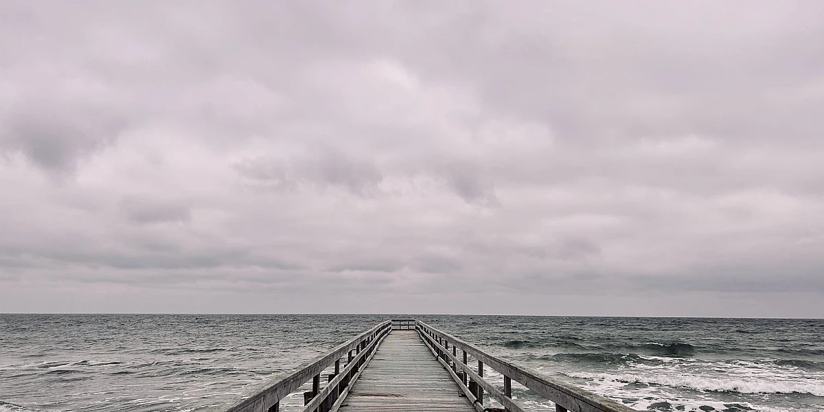 Im unteren Drittel des Bildes ragt eine Holzbrücke in das wilde grüne Meer. Oberhalb des Horizonts dichte Wolken. Alles ist grau.