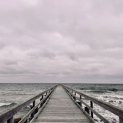 Im unteren Drittel des Bildes ragt eine Holzbrücke in das wilde grüne Meer. Oberhalb des Horizonts dichte Wolken. Alles ist grau.