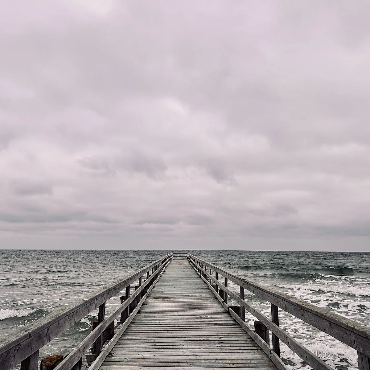 Im unteren Drittel des Bildes ragt eine Holzbrücke in das wilde grüne Meer. Oberhalb des Horizonts dichte Wolken. Alles ist grau.