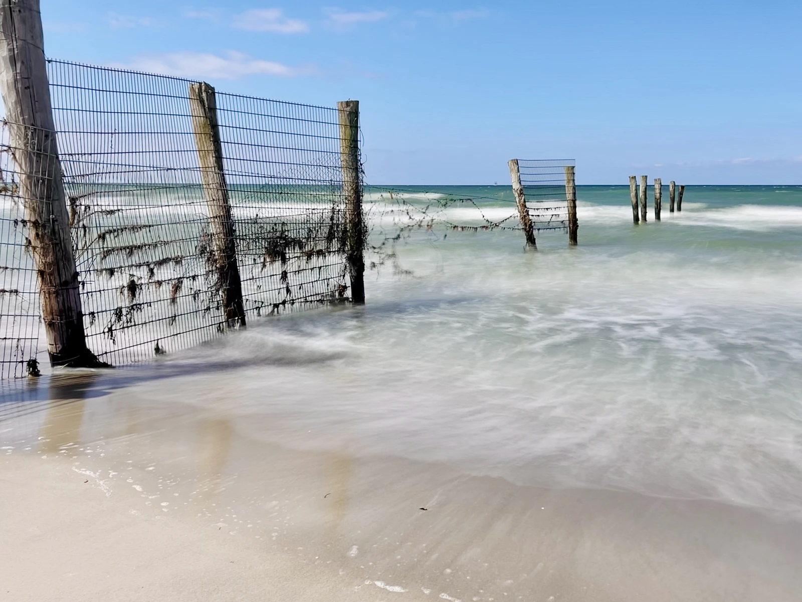 Ein Zaun, der ins Meer ragt ist umgeben vom weich gezeichneten Meer. Der Himmel ist blau.