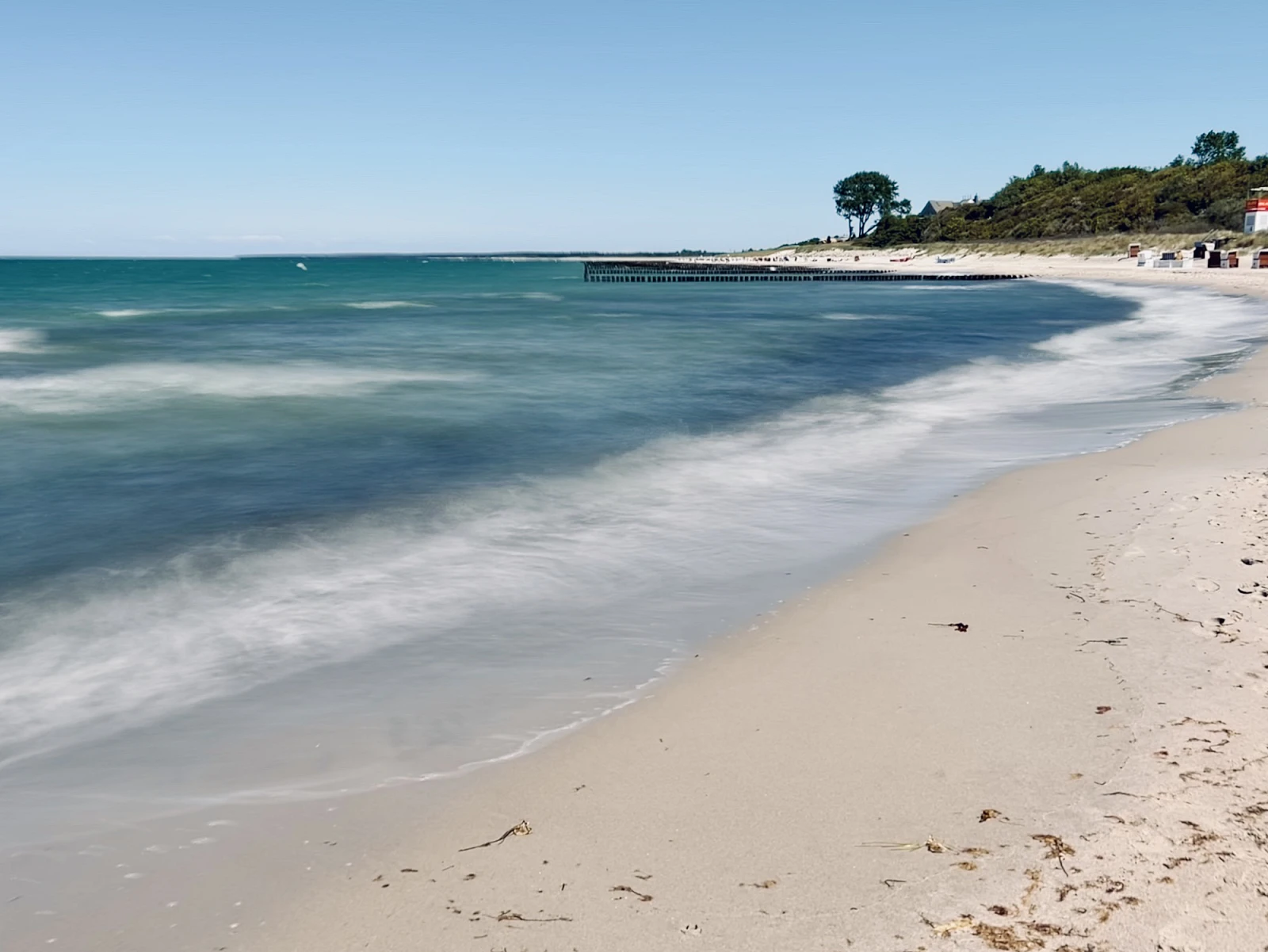 Langzeitaufnahme der Ostsee im linken Teil des Bildes, rechts Sandstrand, Bäume und Büsche am Horizont.