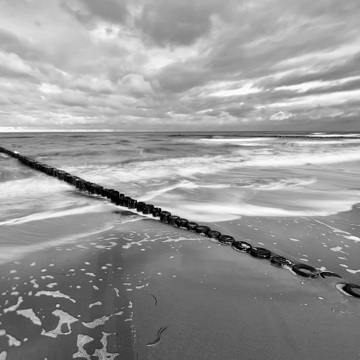 Schwarzweiß-Aufnahme der Ostsee. Eine Holzbuhne zieht sich diagonal von links oben nach rechts unten durchs Bild.