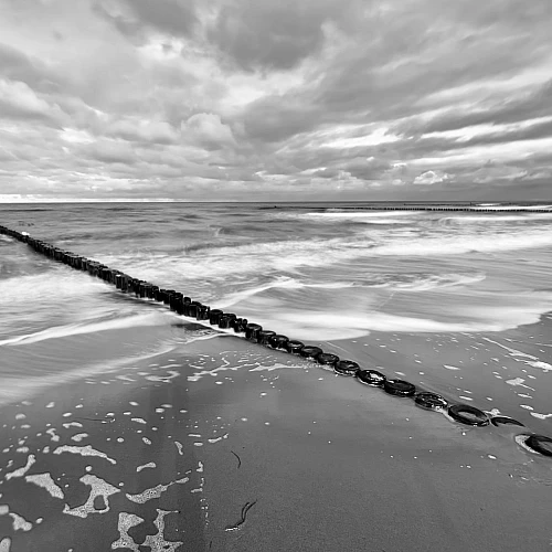 Schwarzweiß-Aufnahme der Ostsee. Eine Holzbuhne zieht sich diagonal von links oben nach rechts unten durchs Bild.