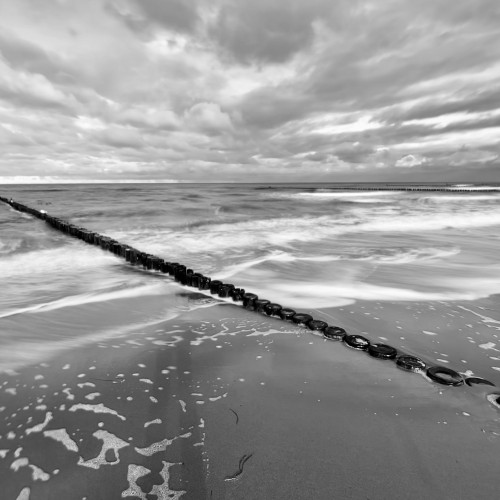 Schwarzweiß-Aufnahme der Ostsee. Eine Holzbuhne zieht sich diagonal von links oben nach rechts unten durchs Bild.