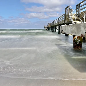 Die Seebrücke von Rerik ist am rechten Rand angeschnitten. Sie ist umgeben vom durch die Langzeitbelichtung weich gewaschen Meer. Der Himmel ist blau mit kleinen weißen Wolken.