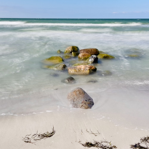 Im Mittelpunkt liegen mehrere große Steinen am Strand und in der Ostsee. Das Wasser drumherum ist durch Langzeitbelichtung verwaschen.