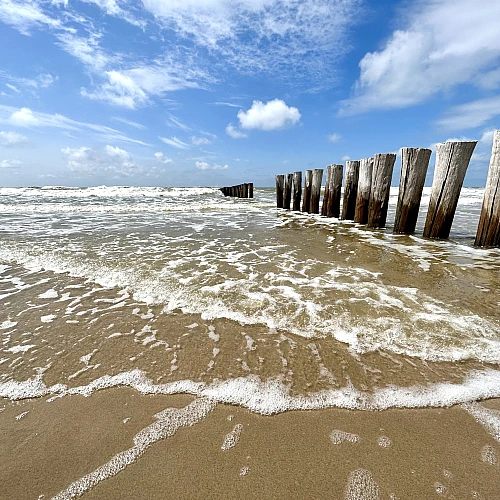 Rechts zieht sich vom Horizon zum Bildrand eine Reihe Holzbuhnen. Das Bild ist horizontal geteilt in ein Drittel Himmel, zwei Drittel Meer mit leichter auslaufender Brandung.