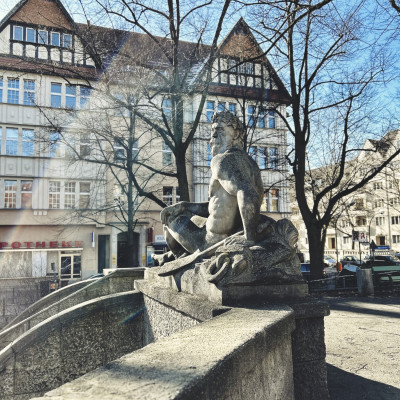 Die Statue eines sitzenden Mannes im Vordergrund, im Hintergrun ein Haus mit zwei spitzen Giebeln.