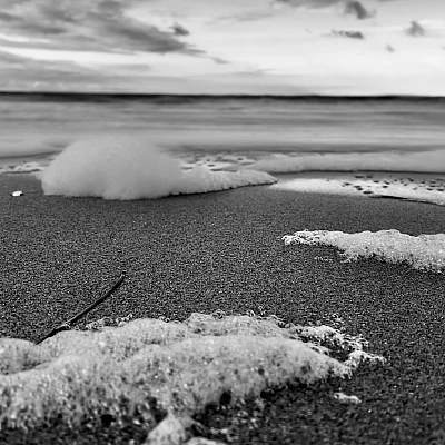 Schwarzweiß-Aufnahme der Ostsee. Im Vordergrund sind Häufchen von Meerschaum auf dem Strand zu sehen.