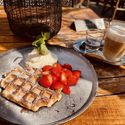 Auf einem Holztisch in warmen Sonnenlicht stehen ein Teller und eine Kaffeetasse mit kleinem Wasserglas. Auf dem Teller eine Belgische Waffel mit Erdbeeren und Schlagsahne.
