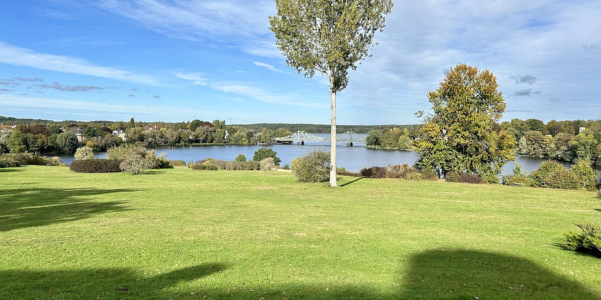 Das Bild zeigt eine malerische Landschaft mit einer weitläufigen, grünen Wiese im Vordergrund. Auf der Wiese stehen zwei Bäume, einer mit grünen Blättern und der andere mit herbstlich gefärbten Blättern. Im Hintergrund sieht man einen ruhigen See, umgeben von Bäumen und Sträuchern. Jenseits des Sees sind Häuser und eine Brücke zu erkennen. Der Himmel ist blau mit einigen weißen Wolken. Es sieht nach einem sonnigen Tag aus.