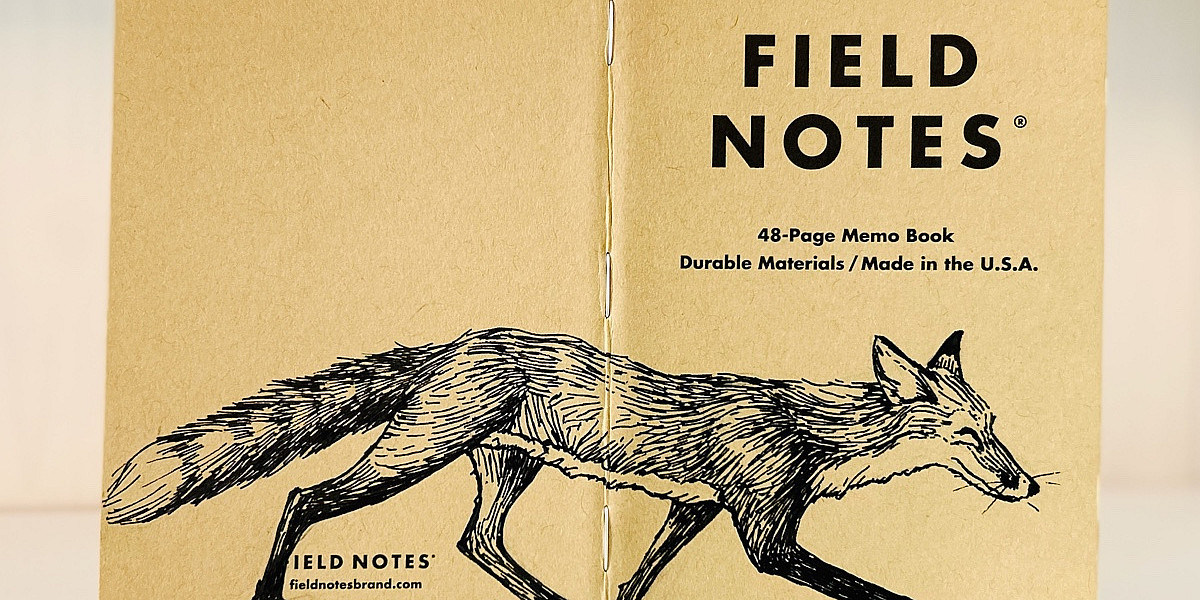Auf dem aufgeklappten Rücken eines Field Notes Heftes ist eine schwarze Tintezeichnung eines Fuchses zu sehen. Der sehr schlanke Fuchs läuft von links nach rechts.