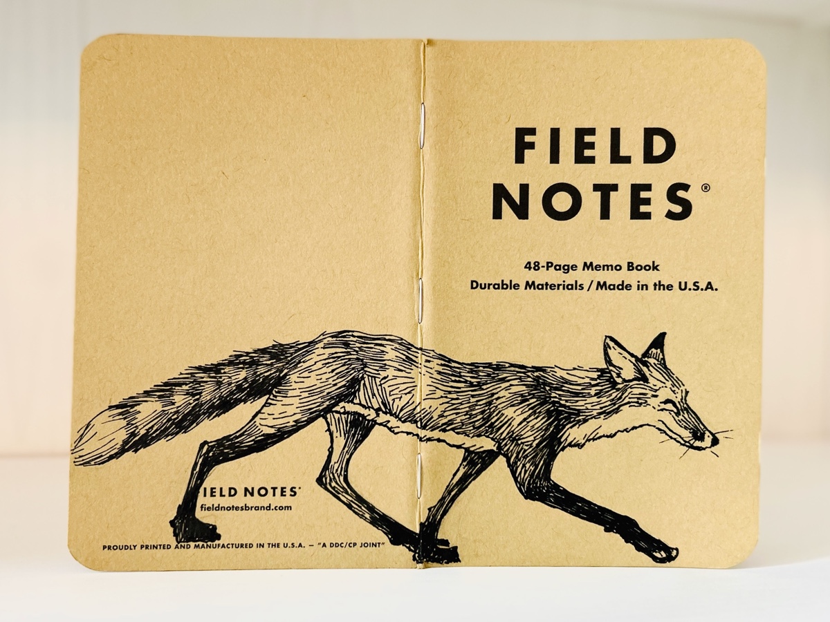 Auf dem aufgeklappten Rücken eines Field Notes Heftes ist eine schwarze Tintezeichnung eines Fuchses zu sehen. Der sehr schlanke Fuchs läuft von links nach rechts.