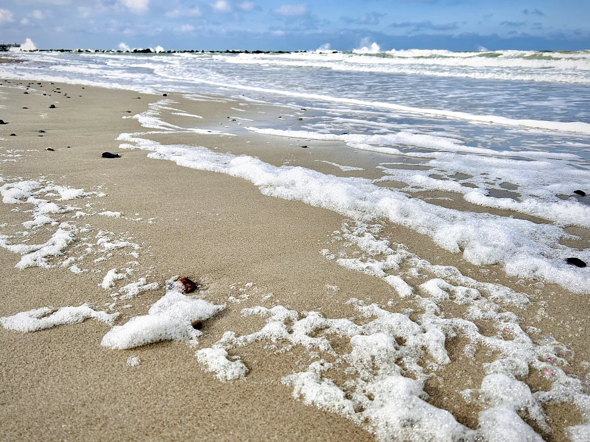 Weißer Schaum und schäumende Brandung am Sandstrand. Im Hinterrund sind brechende Wellen. Ein schmaler Streifen blauen Himmels mit weißen Wolken am oberen Bildrand.