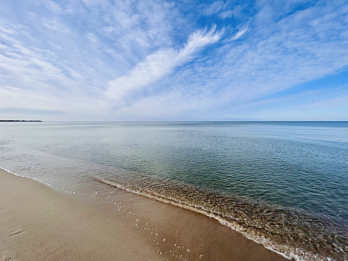 Das Bild ist zweigeteilt. Über dem Horizon der blaue Himmel mit weißen Wolkenstreifen. Im unteren Teil des Bildes das stille blaugrüne Meer und ganz links etwas Sandstrand.