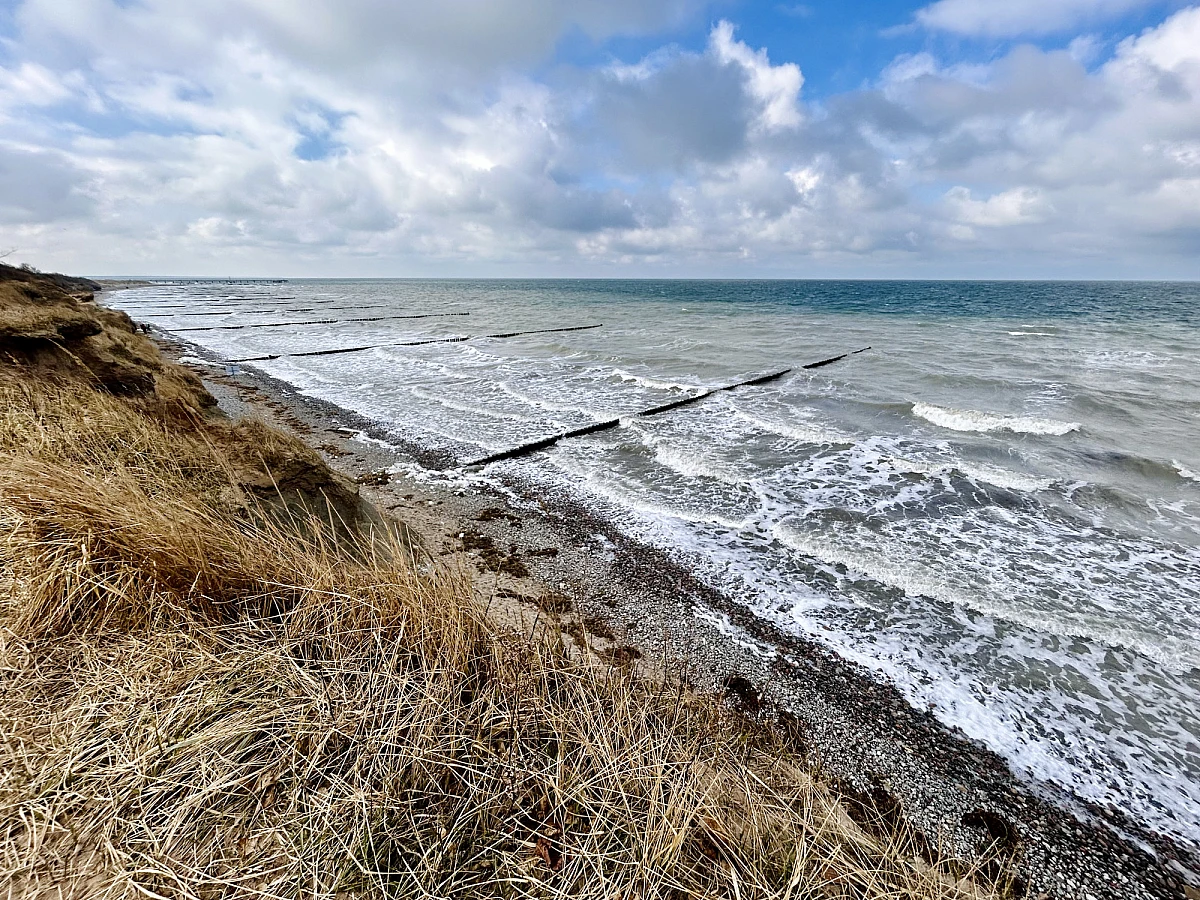 Blick vom Kliff mit vertrocknetem Gras auf das schäumende grüngraue Meer mit Stehlen. Der blaue Himmel ist wolkenverhangen.