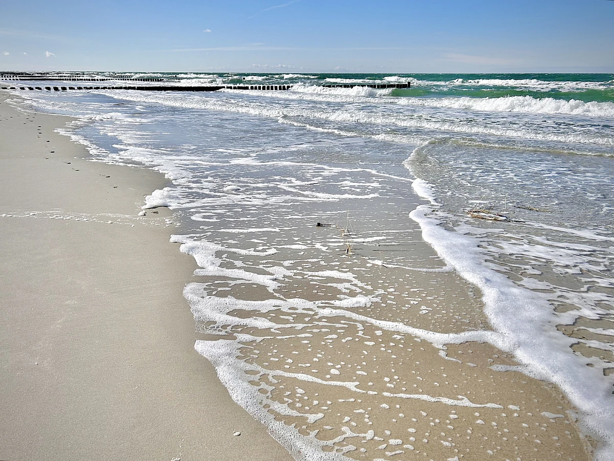 Weißer Schaum und schäumende Brandung am Sandstrand. Im Hinterrund sind brechende Wellen. Ein schmaler Streifen blauen Himmels mit weißen Wolken am oberen Bildrand.