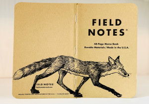 Auf dem aufgeklappten Rücken eines Field Notes Heftes ist eine schwarze Tintezeichnung eines Fuchses zu sehen. Der sehr schlanke Fuchs läuft von links nach rechts.