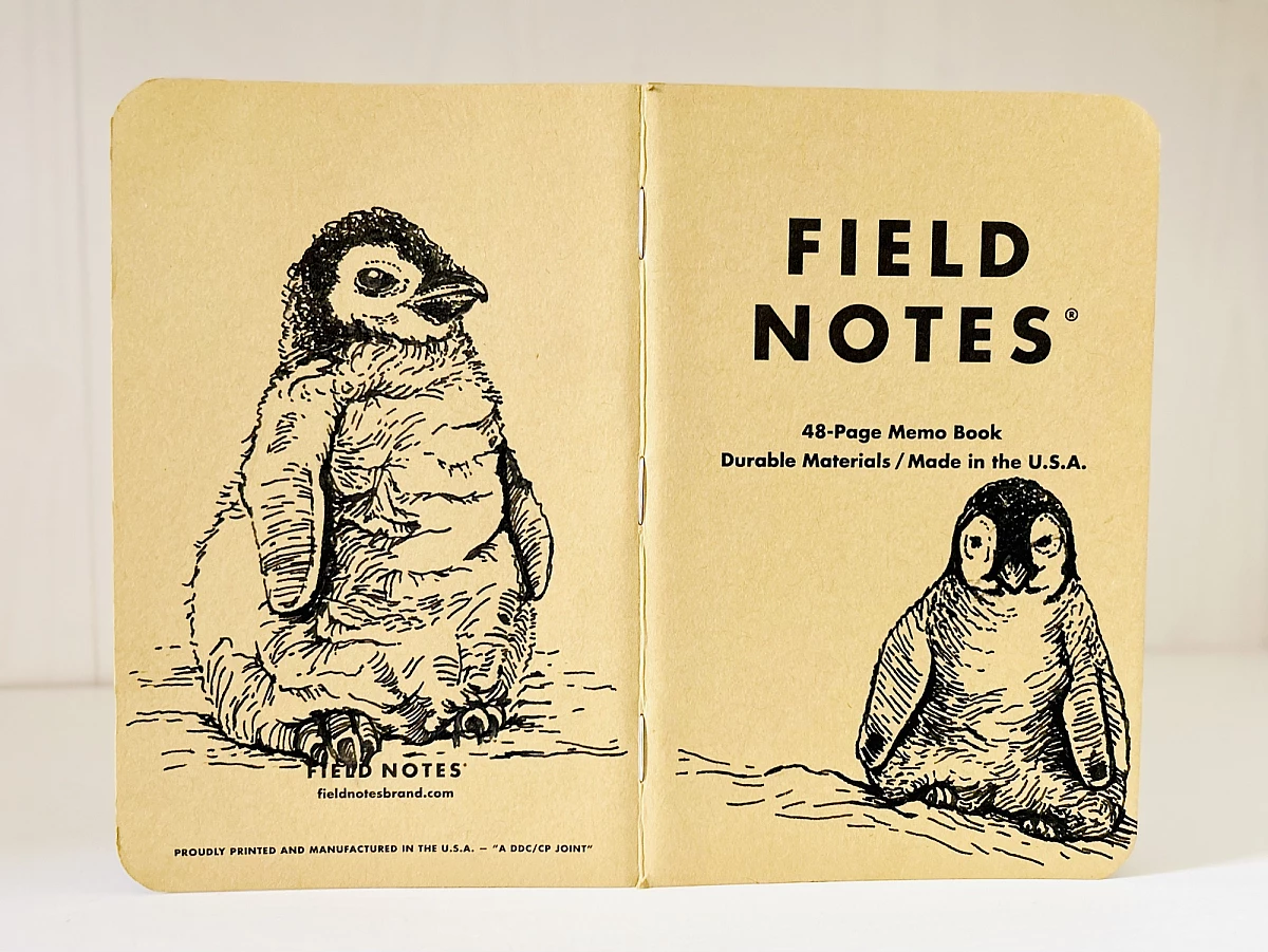 Foto eines aufgeklappten Field Notes Heftes mit Kraft-Papier-Deckel. Auf Rücken und Cover befindet sich jeweils eine schwarze Zeichnung eines sitzenden Baby Pinguins.