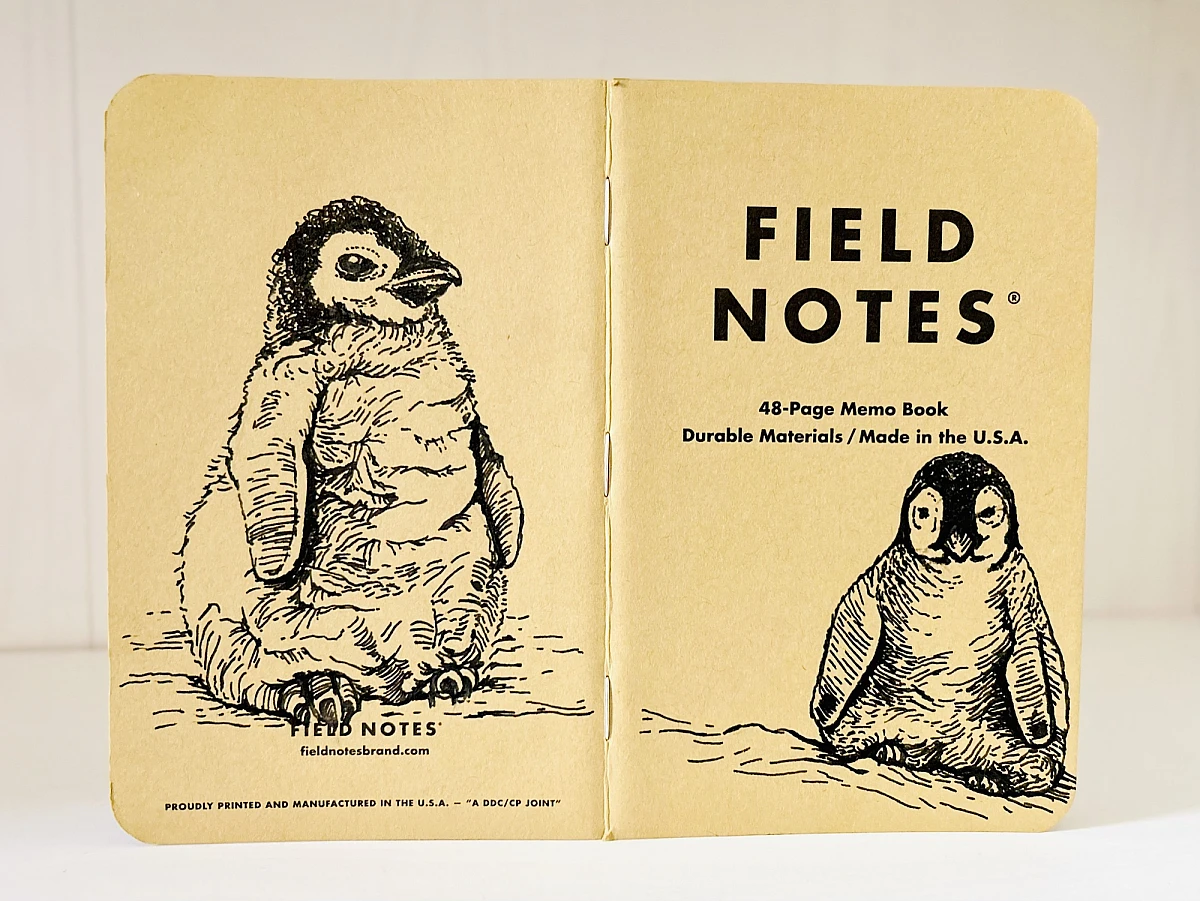 Foto eines aufgeklappten Field Notes Heftes mit Kraft-Papier-Deckel. Auf Rücken und Cover befindet sich jeweils eine schwarze Zeichnung eines sitzenden Baby Pinguins.