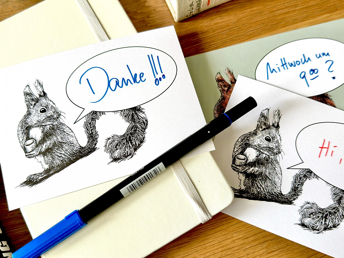 Eine Postkarte mit dem Motiv eines Eichhörnchens und einer Sprechblase und dem handschriftlichen Schriftzug "Happy Monday" liegt zusammen mit einem Füllhalter auf einem Notizbuch auf einem Holztisch.