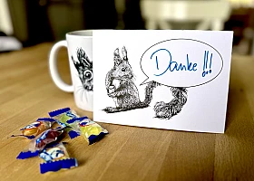 Eine Postkarte mit dem Motiv eines Eichhörnchens mit Sprechblase steht aufrecht gegen eine Tasse gelehnt auf einem Holztisch. Davor ein paar Bonbons. Auf der Karte steht handschriftlich "Danke!!!".