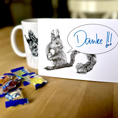 Eine Postkarte mit dem Motiv eines Eichhörnchens mit Sprechblase steht aufrecht gegen eine Tasse gelehnt auf einem Holztisch. Davor ein paar Bonbons. Auf der Karte steht handschriftlich "Danke!!!".