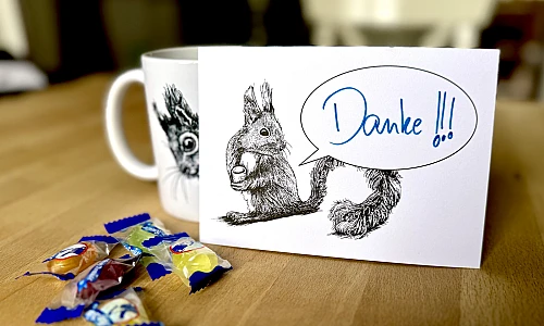 Eine Postkarte mit dem Motiv eines Eichhörnchens mit Sprechblase steht aufrecht gegen eine Tasse gelehnt auf einem Holztisch. Davor ein paar Bonbons. Auf der Karte steht handschriftlich "Danke!!!".