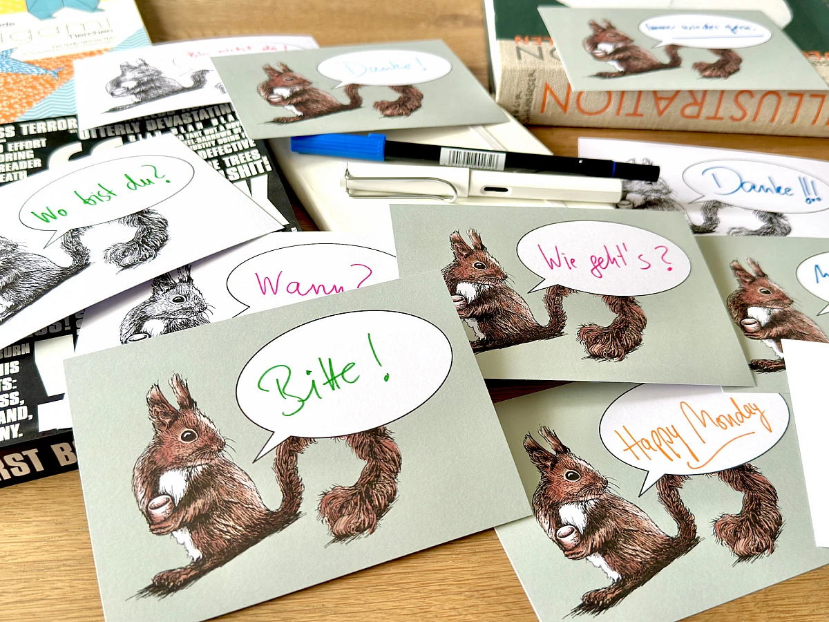 Eine Postkarte mit dem Motiv eines Eichhörnchens und einer Sprechblase und dem handschriftlichen Schriftzug "Happy Monday" liegt zusammen mit einem Füllhalter auf einem Notizbuch auf einem Holztisch.