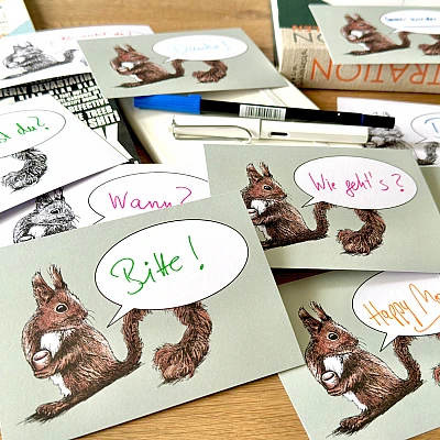 Verschiedene Postkarten mit mit dem Motiv eines Eichhörnchen und einer Sprechblase liegen auf einen Holztisch. In den Sprechblasen finden sich handschriftlich Schriftzüge wie "Bitte!", "Wie geht's?", "Wann?", "Happy Monday"