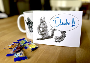 Eine Postkarte mit dem Motiv eines Eichhörnchens mit Sprechblase steht aufrecht gegen eine Tasse gelehnt auf einem Holztisch. Davor ein paar Bonbons. Auf der Karte steht handschriftlich "Danke!!!".