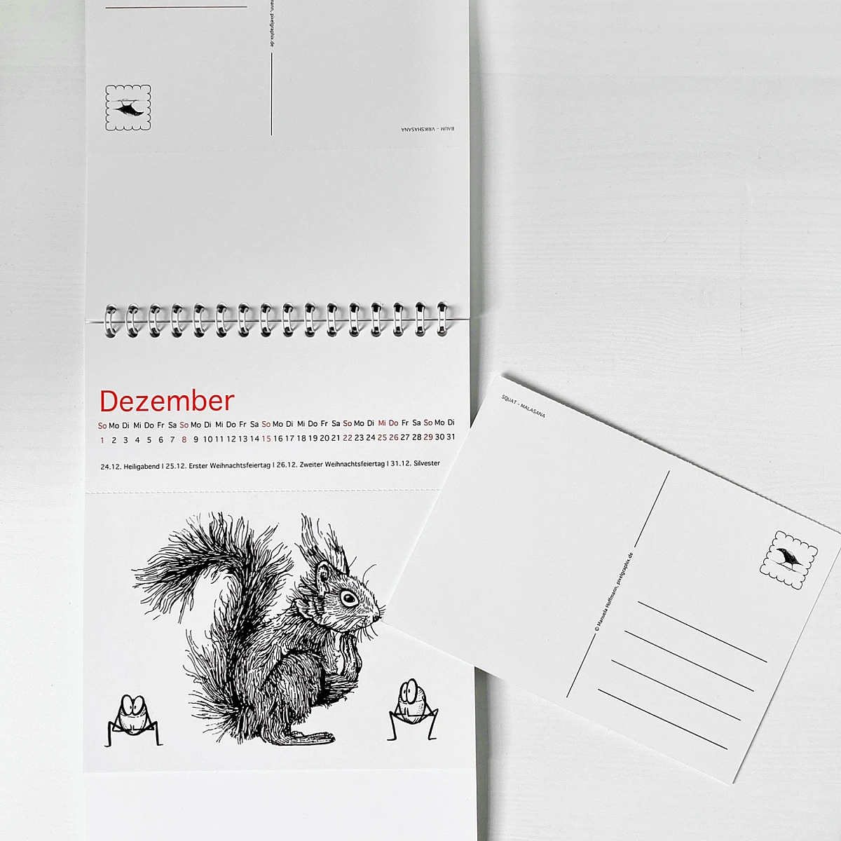Der Kalender Ommmmmmmmmmmm - Yoga für Eichhörnchen & ähnlich veranlagte Menschen - Postkartenkalender 2024  steht seitlich aufgeklappt auf einer weißen Fläche. Auf dem Cover ist ein Eichhörnchen zu sehen, das die Position des herabschauenden Hundes praktiziert.  Fünf Haselnüsse praktizieren ebenfalls Yoga.
