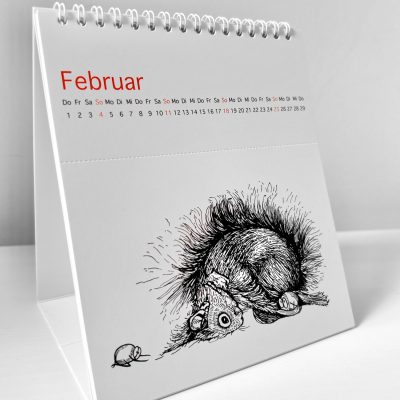 Der Kalender steht aufgeklappt auf einer weißen Fläche. Es ist der Monat Februar zu sehen. Es ist eine Illustration zu sehen, auf der ein Eichhörnchen und zwei Haselnüsse in der Kindspose verweilen.