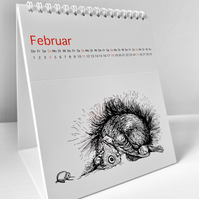 Der Kalender steht aufgeklappt auf einer weißen Fläche. Es ist der Monat Februar zu sehen. Es ist eine Illustration zu sehen, auf der ein Eichhörnchen und zwei Haselnüsse in der Kindspose verweilen.