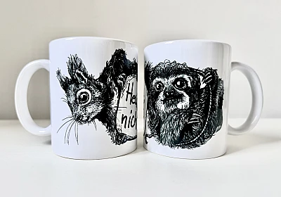 Zwei weiße Keramiktassen stehen nebeneinander. Auf der linken Tasse die schwarze Zeichnung eines Eichhörnchenkopfes, auf der rechten die schwarze Zeichnung eines Faultiers.